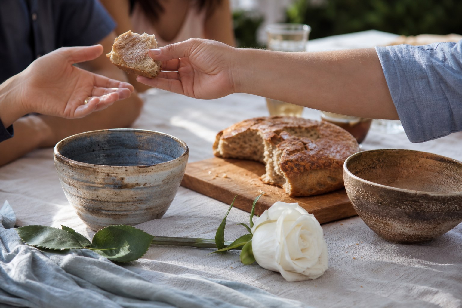 A hand shares bread across a communal table with a white rose and two handmade bowls