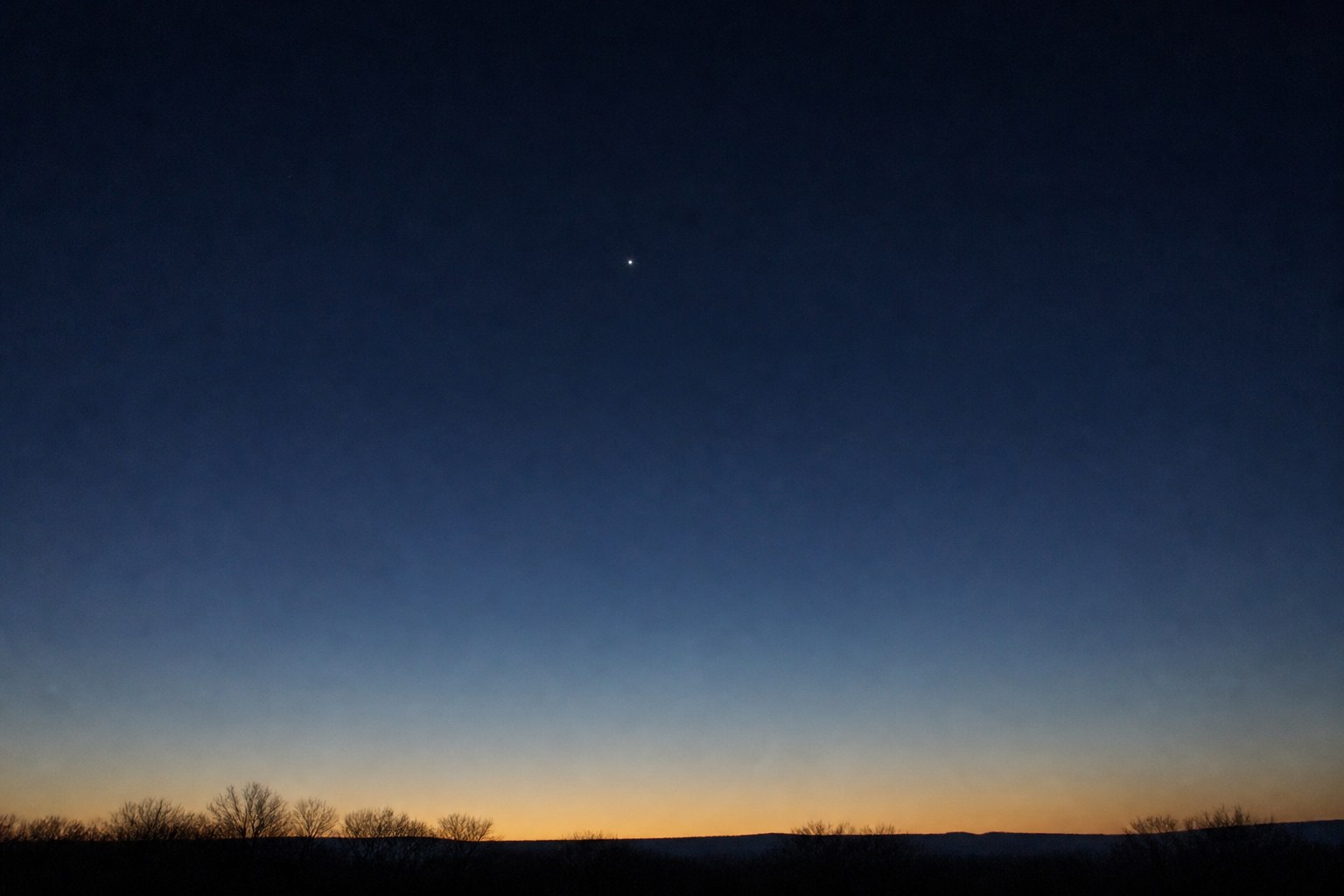 The pre-dawn eastern sky graded from indigo to a thin band of gold at the horizon, with a single bright point of light above a low silhouette of bare trees and a distant ridge