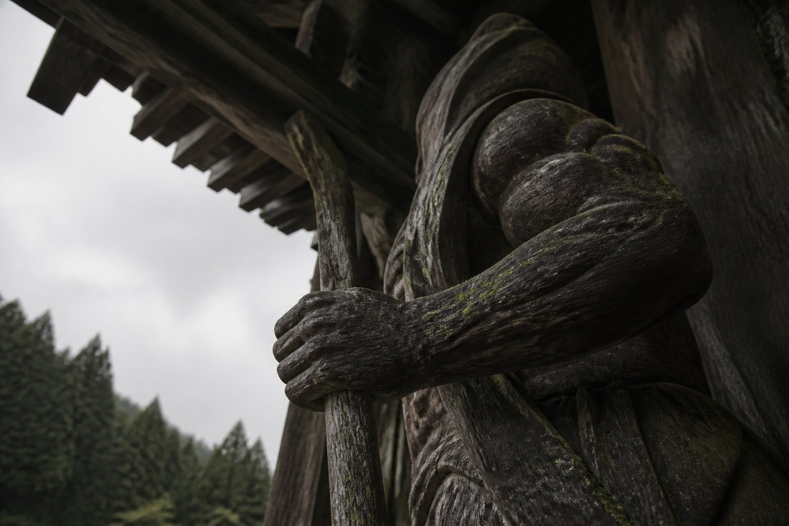 A weathered carved wooden temple gate guardian figure seen from below against an overcast sky, moss growing on the timber
