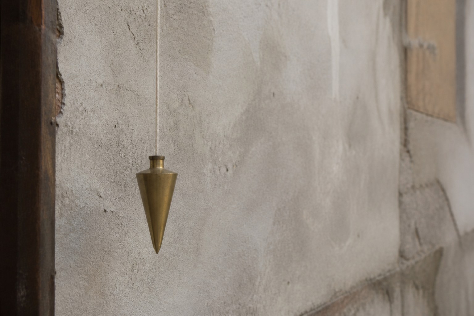 A brass plumb bob hanging still on its cord before a rough plastered wall