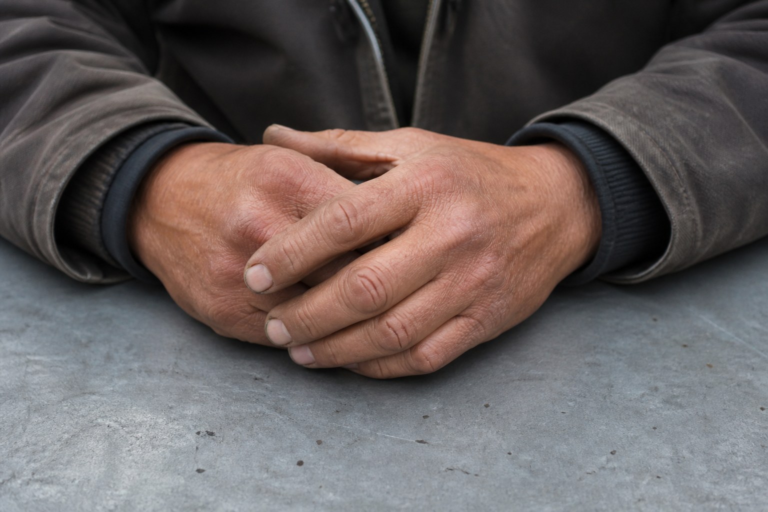 Two weathered hands clasped loosely over a plain tabletop
