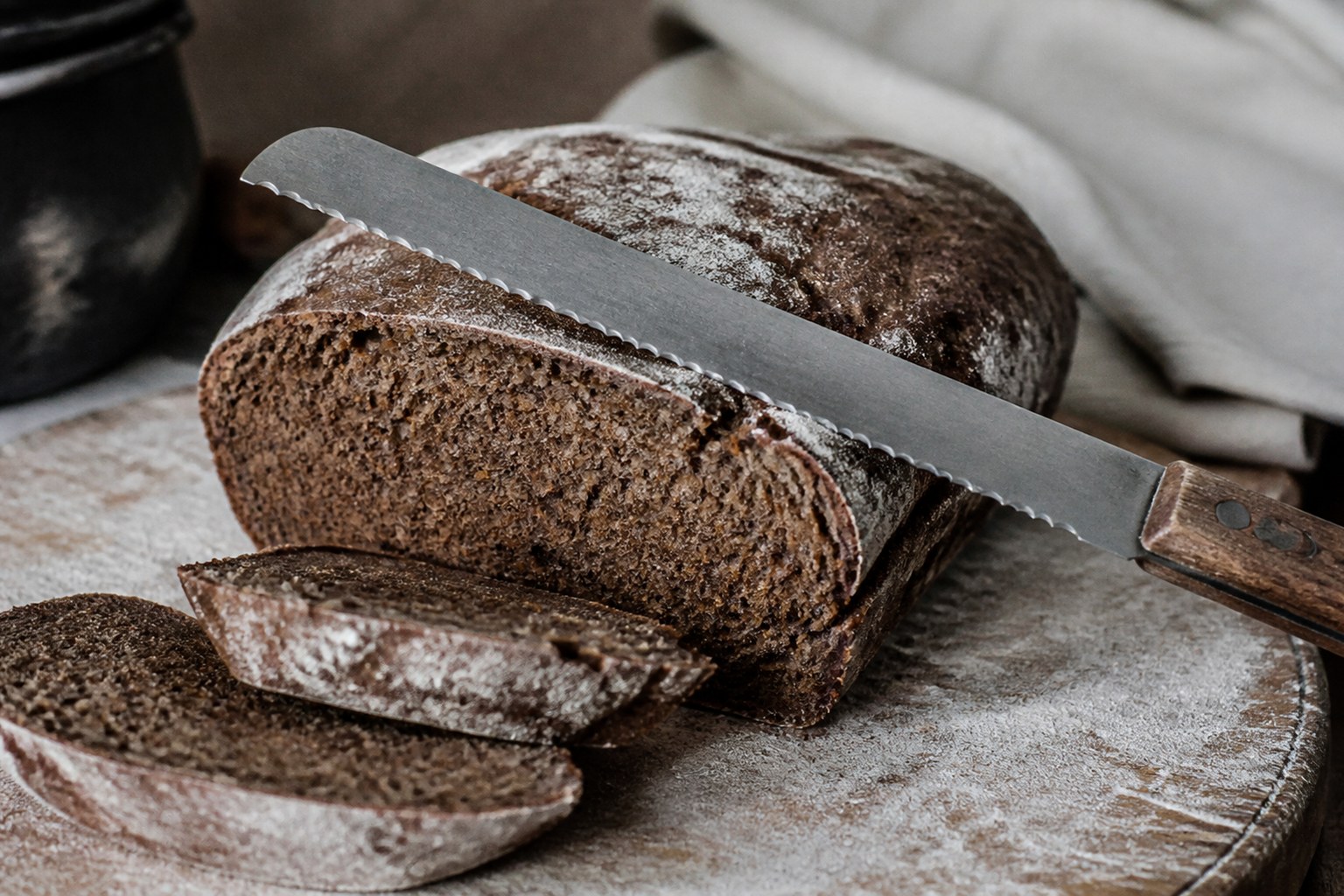 A serrated knife resting on the cut face of a dark loaf on a floured board