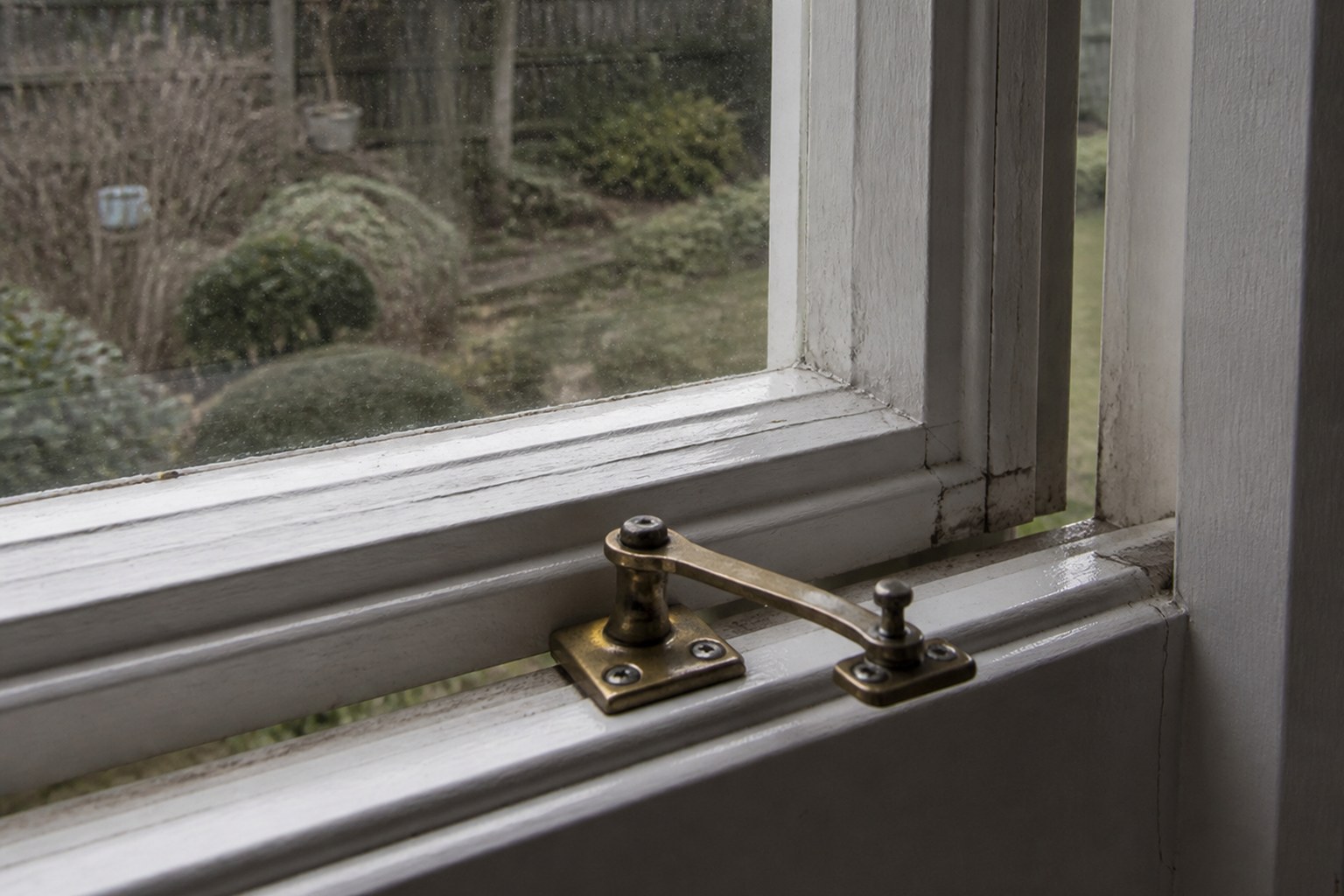 A brass casement catch holding a window open onto a grey garden