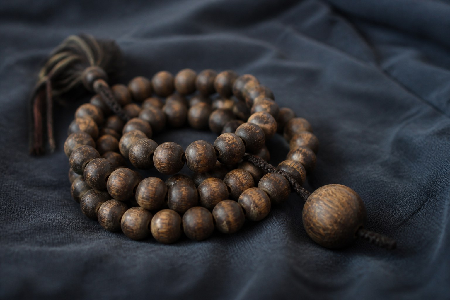A worn wooden prayer bead mala coiled on a dark cloth with one larger bead standing slightly apart from the loop