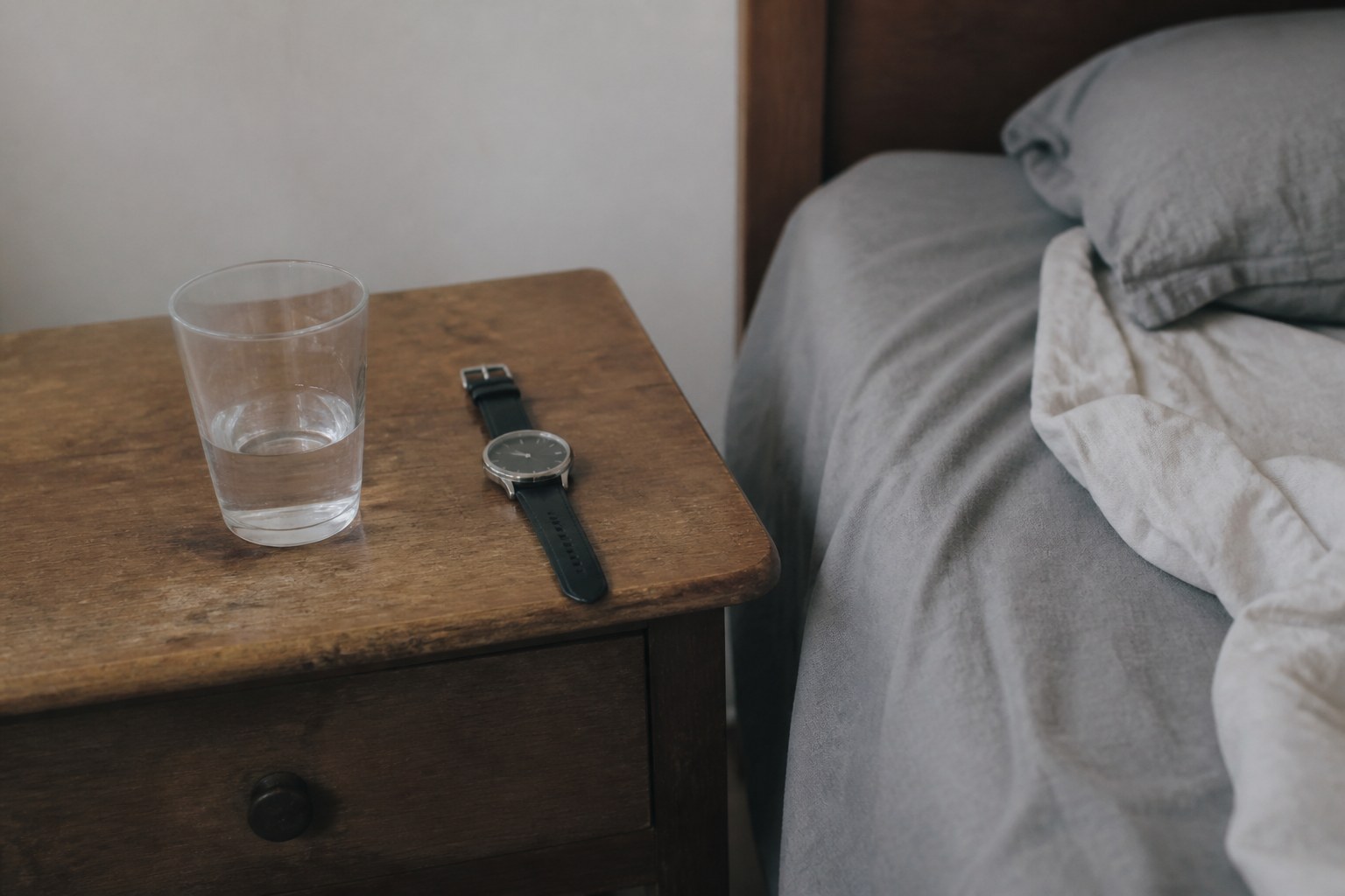 A bedside table at first light with a half-drunk glass of water and a wristwatch beside an unmade bed