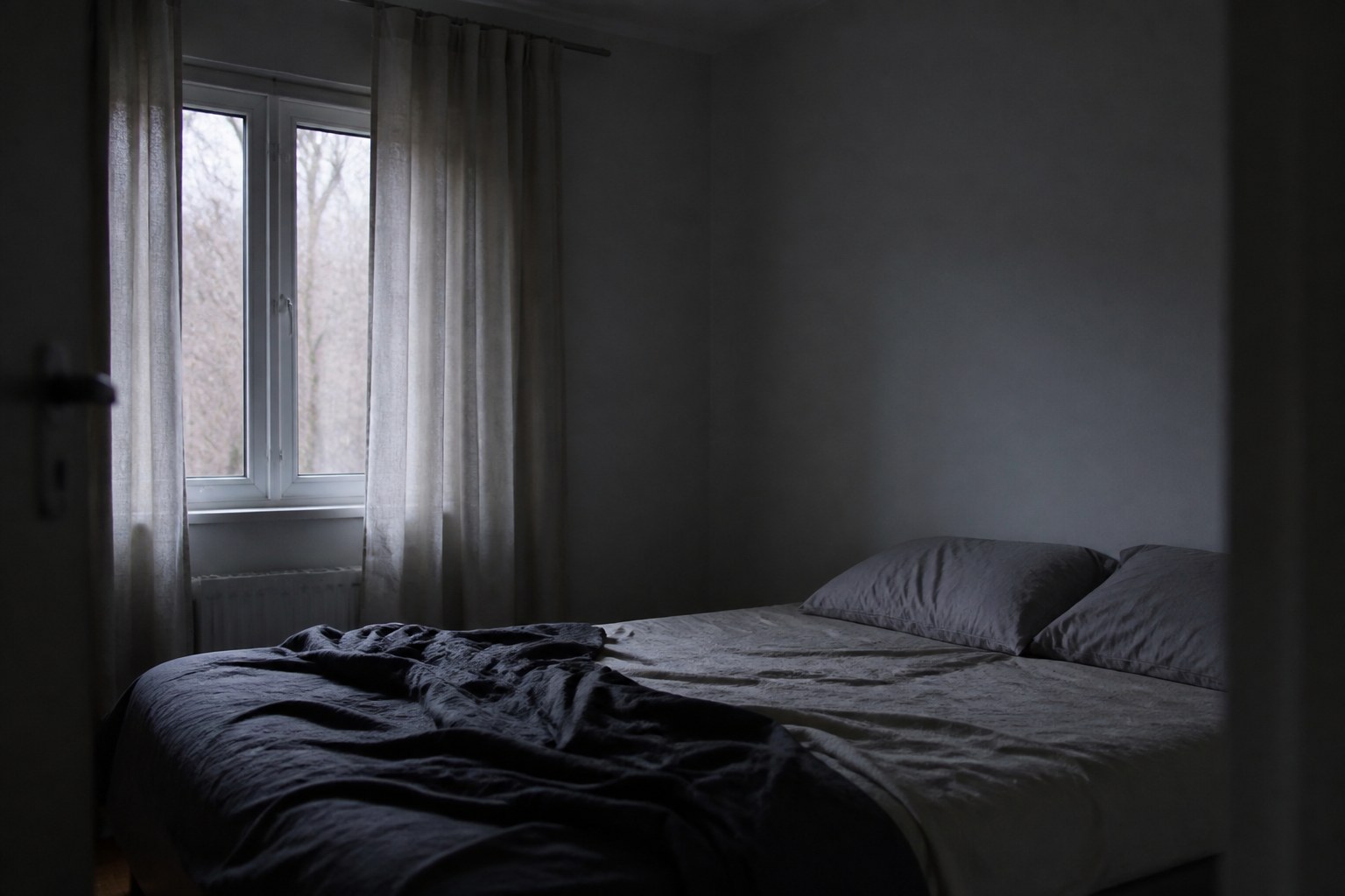 A bedroom in grey pre-dawn light with the bed unmade, the duvet pushed aside, and thin curtains half drawn at the window