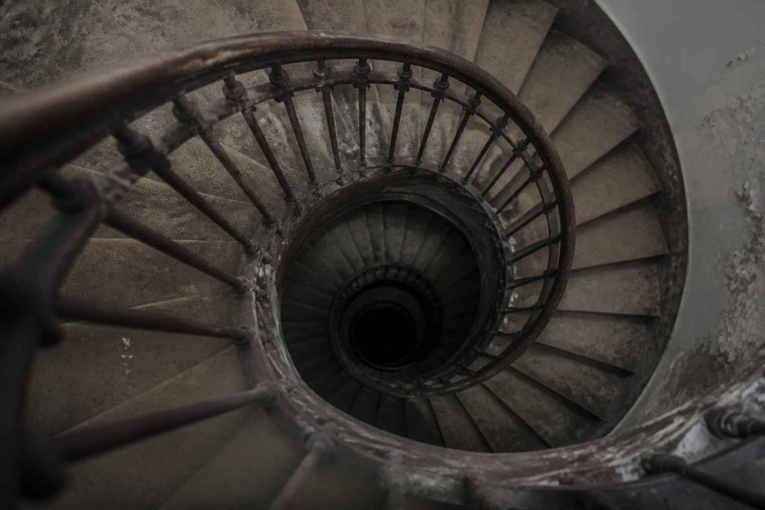 The open well of a stone spiral staircase seen from the top floor, worn steps and an iron balustrade curving down into shadow