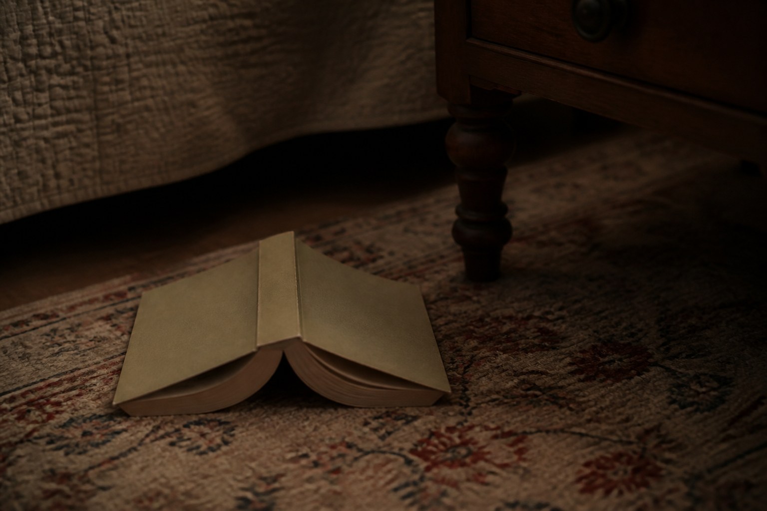 A paperback lying face down and splayed open on a rug beside the leg of a bedside table, in low lamplight