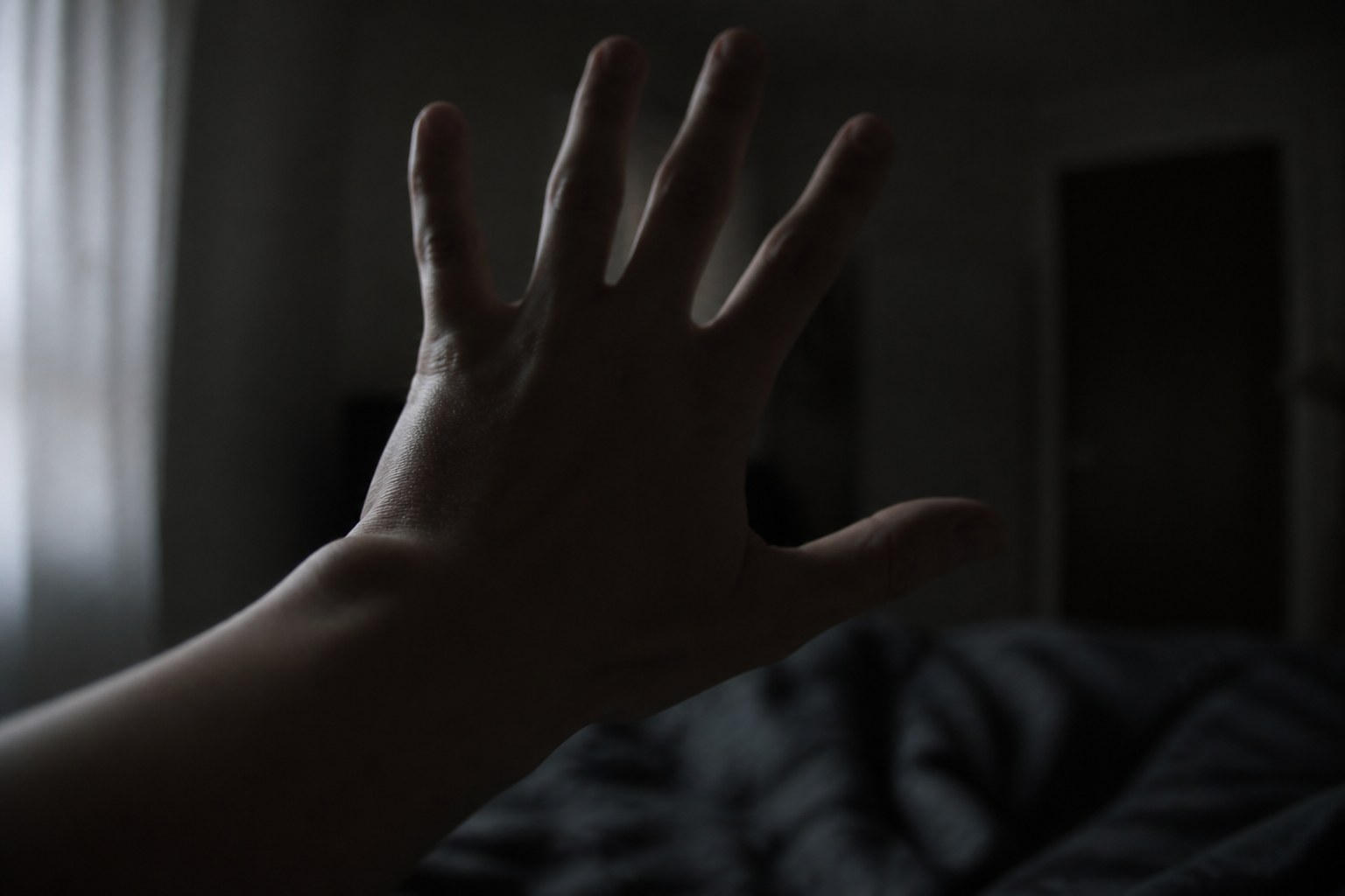 A hand held up close in a dim bedroom, fingers spread and slightly blurred, the bed dark below