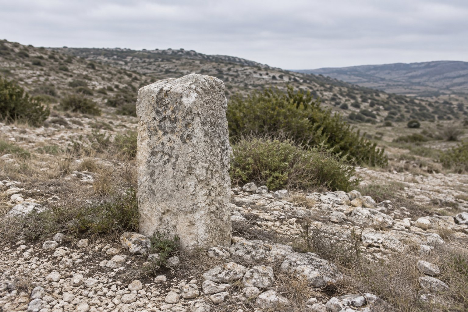 A rough-cut limestone boundary marker standing alone in dry, stony hill country under flat overcast light