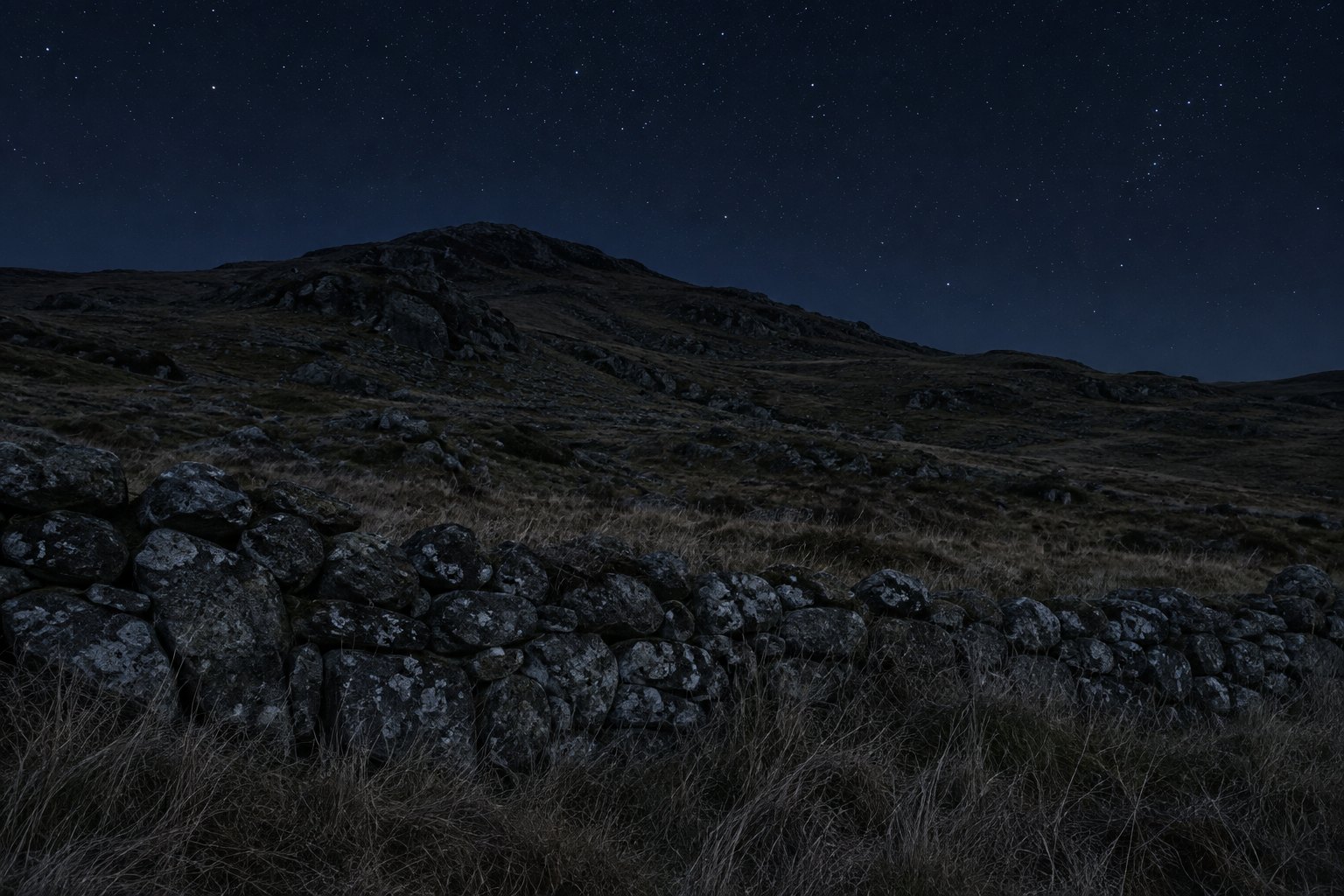 A dark hillside at night under a clear sky, with a low stone wall in the foreground and no light source visible