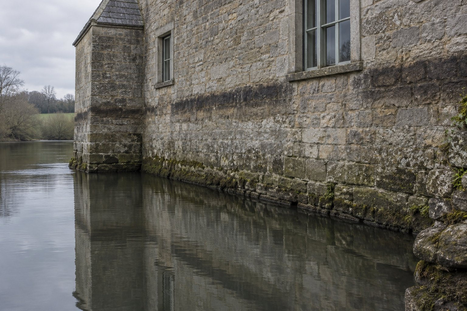 A stone building wall rising from still water, with a dark horizontal high-water stain running across the stonework well above the present waterline