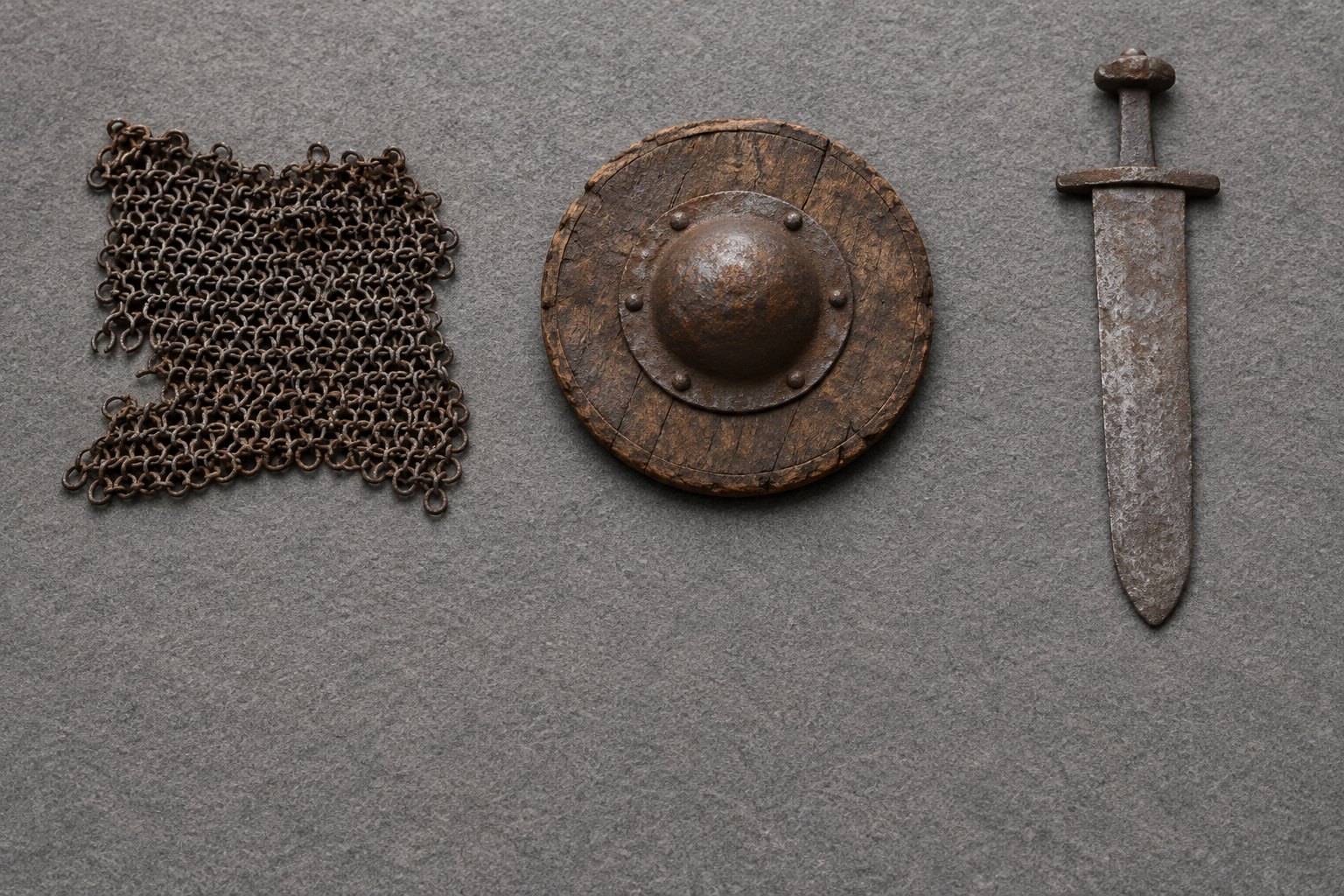 A square of riveted iron mail, a small round wooden shield with an iron boss, and a short corroded iron sword laid apart in a row on grey felt
