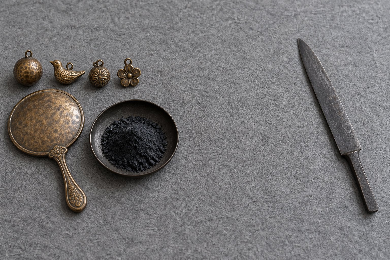 Small hammered bronze ornaments, a bronze hand mirror and a dish of dark powdered pigment grouped at one side of a grey surface, with a single plain iron blade laid well apart at the other