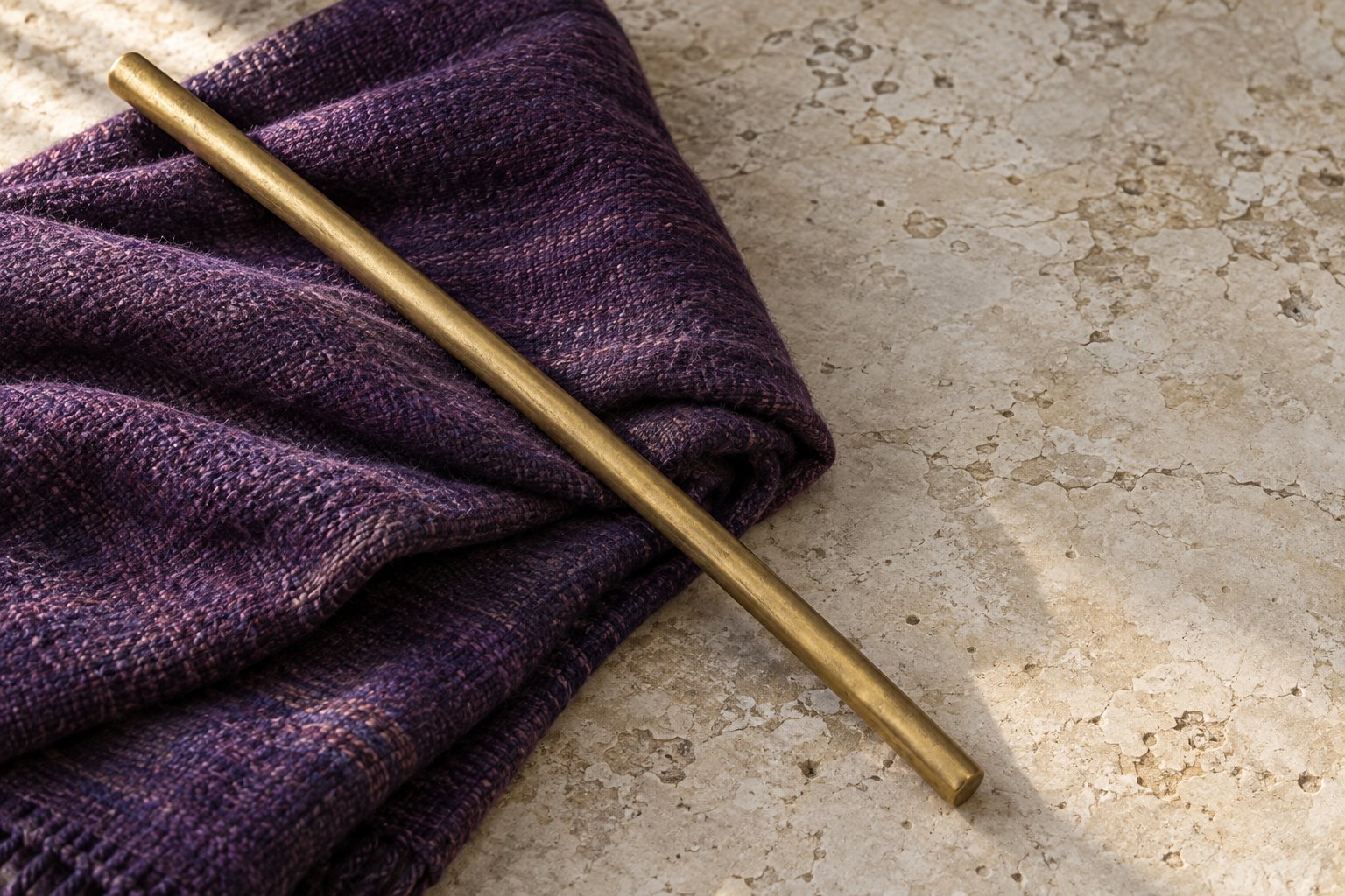 A folded length of deep purple cloth and a plain gold-coloured rod laid on pale stone in soft daylight