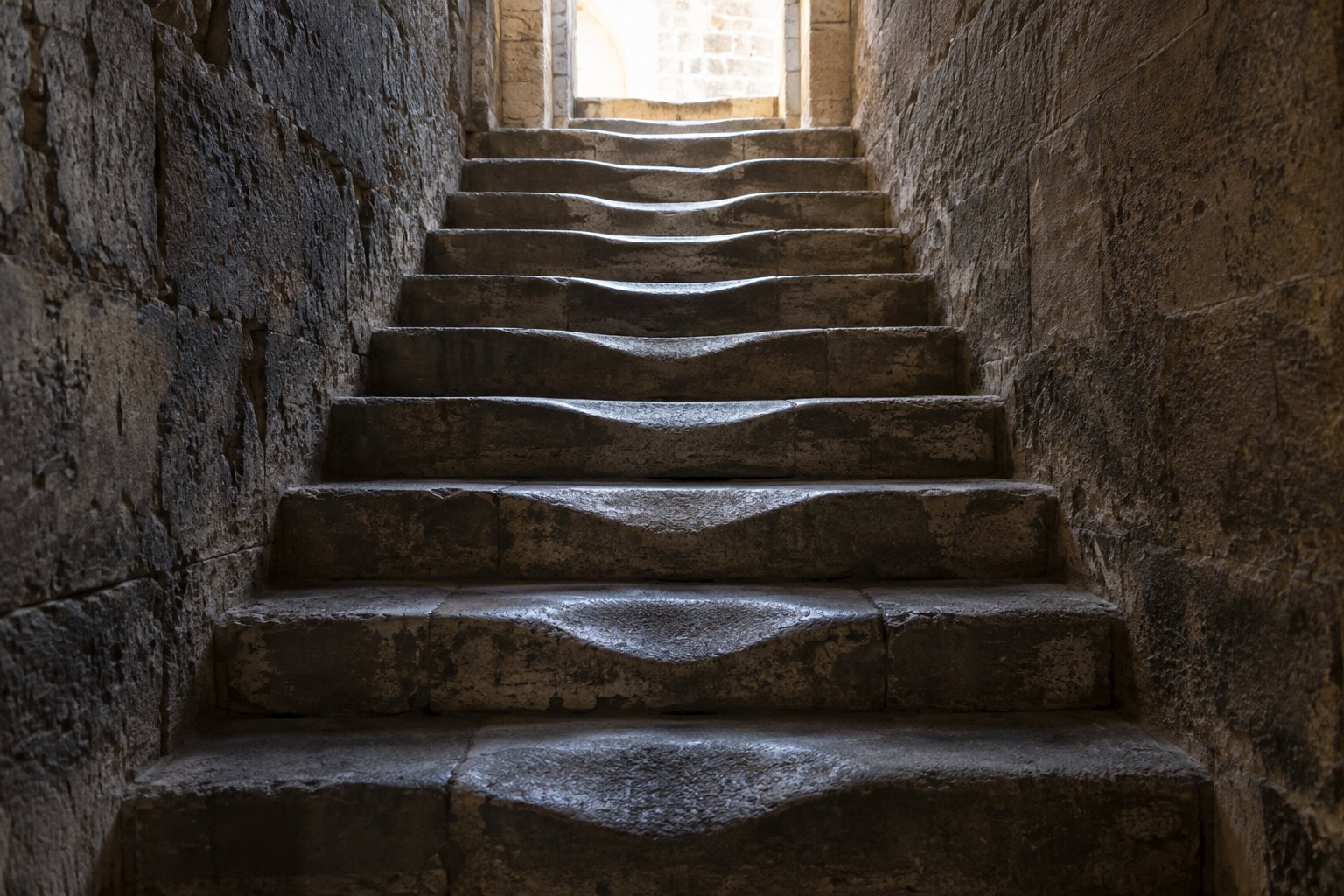 A worn stone stairway inside a dim building, its treads hollowed in the middle by centuries of feet, rising toward a bright opening