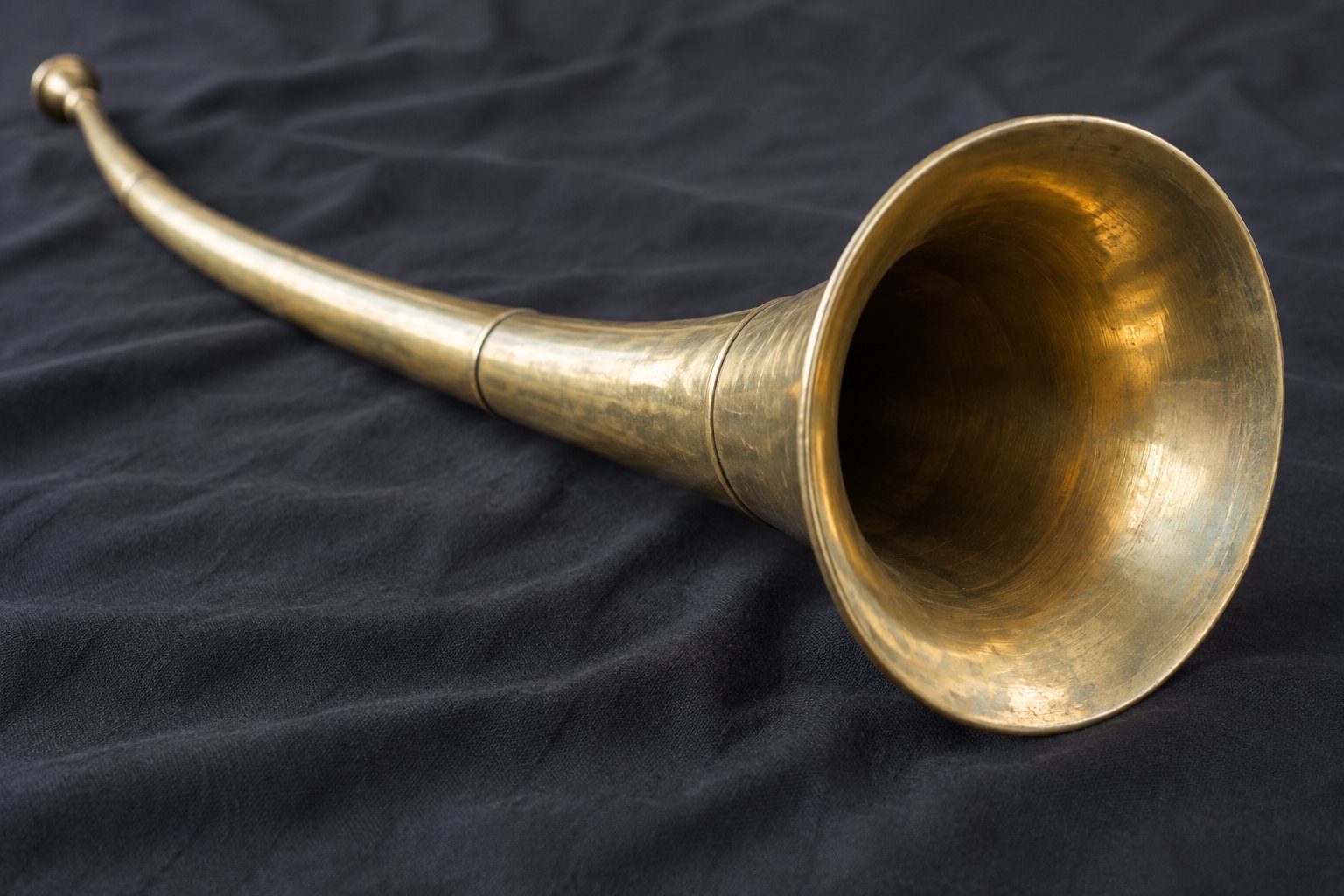 A long curved brass horn resting on a plain dark cloth, its bell turned toward the viewer, lit by soft daylight