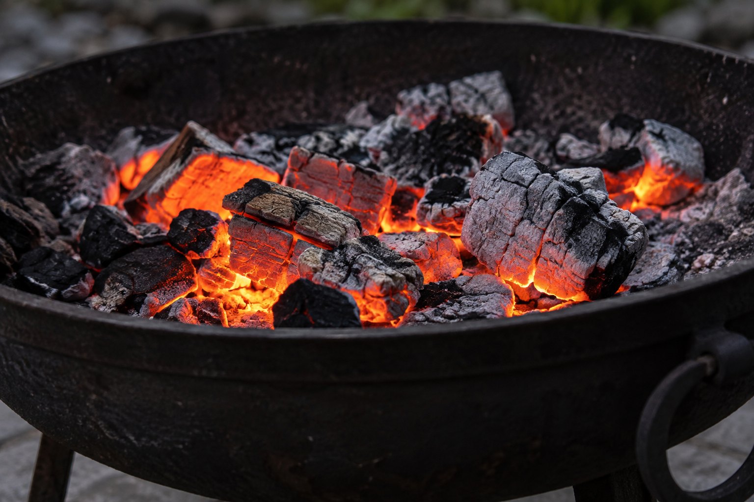 A bed of glowing charcoal embers in a dark iron brazier, some coals bright orange and others crusted grey with ash