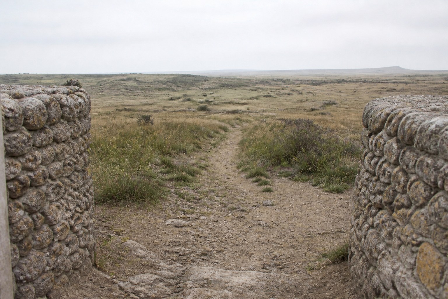 A rough track leading away from a low stone enclosure into empty scrubland