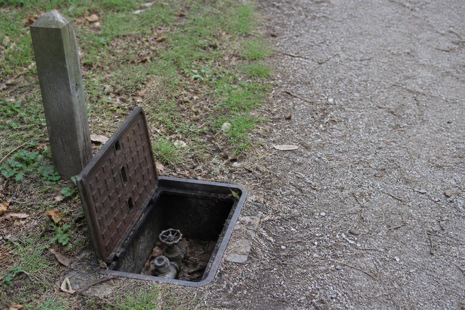 An outdoor stopcock box open in a path with the valve visible and a marker post beside it