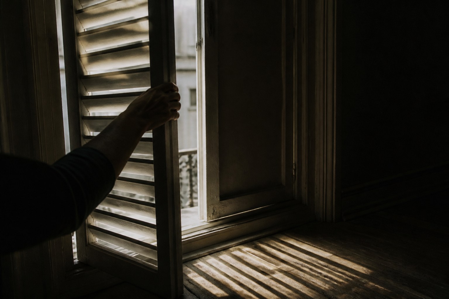 Shutters being opened from inside a dark room, hard daylight cutting across the floor