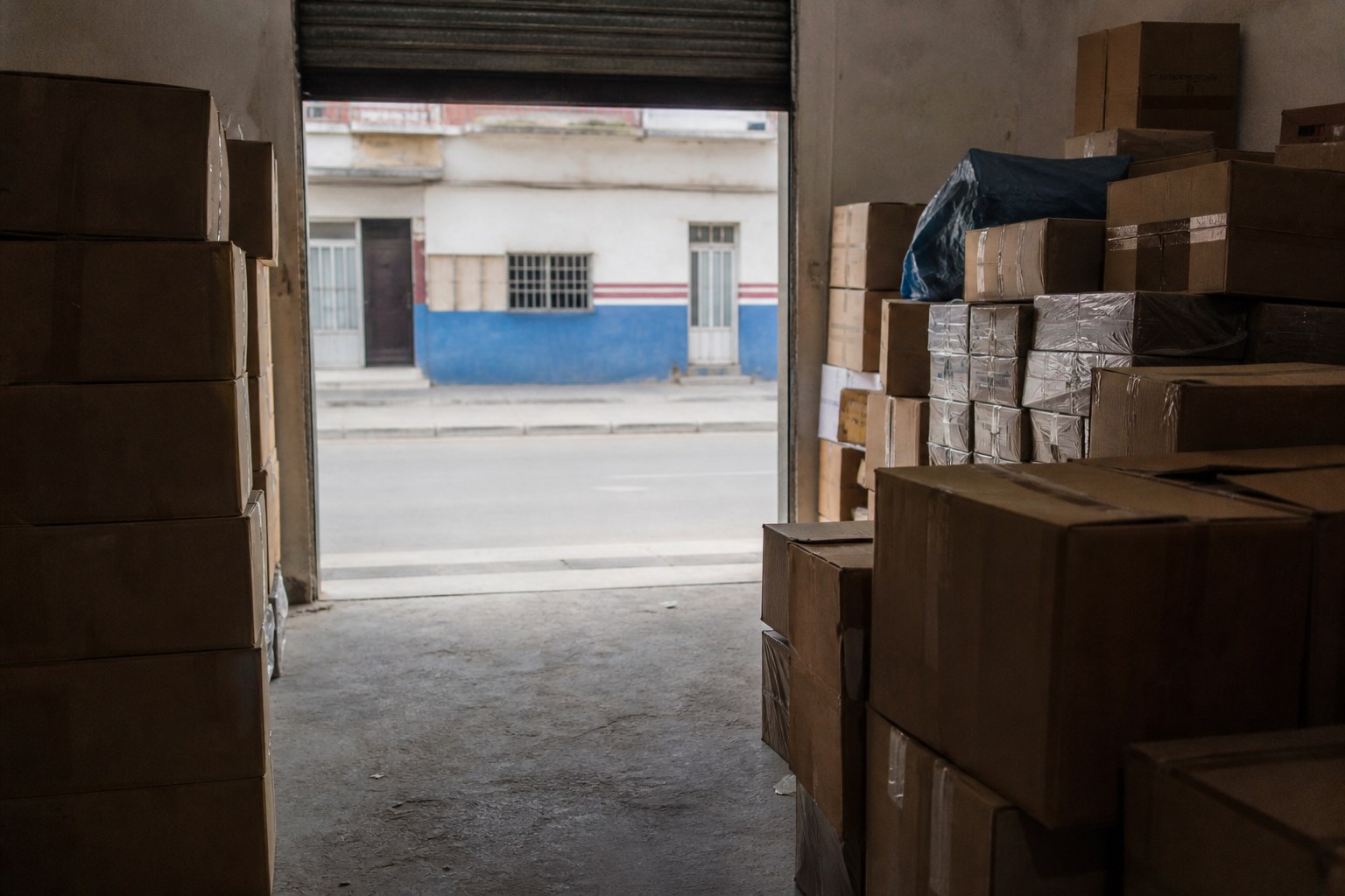 A storeroom stacked with boxed stock, the roller door open onto a street