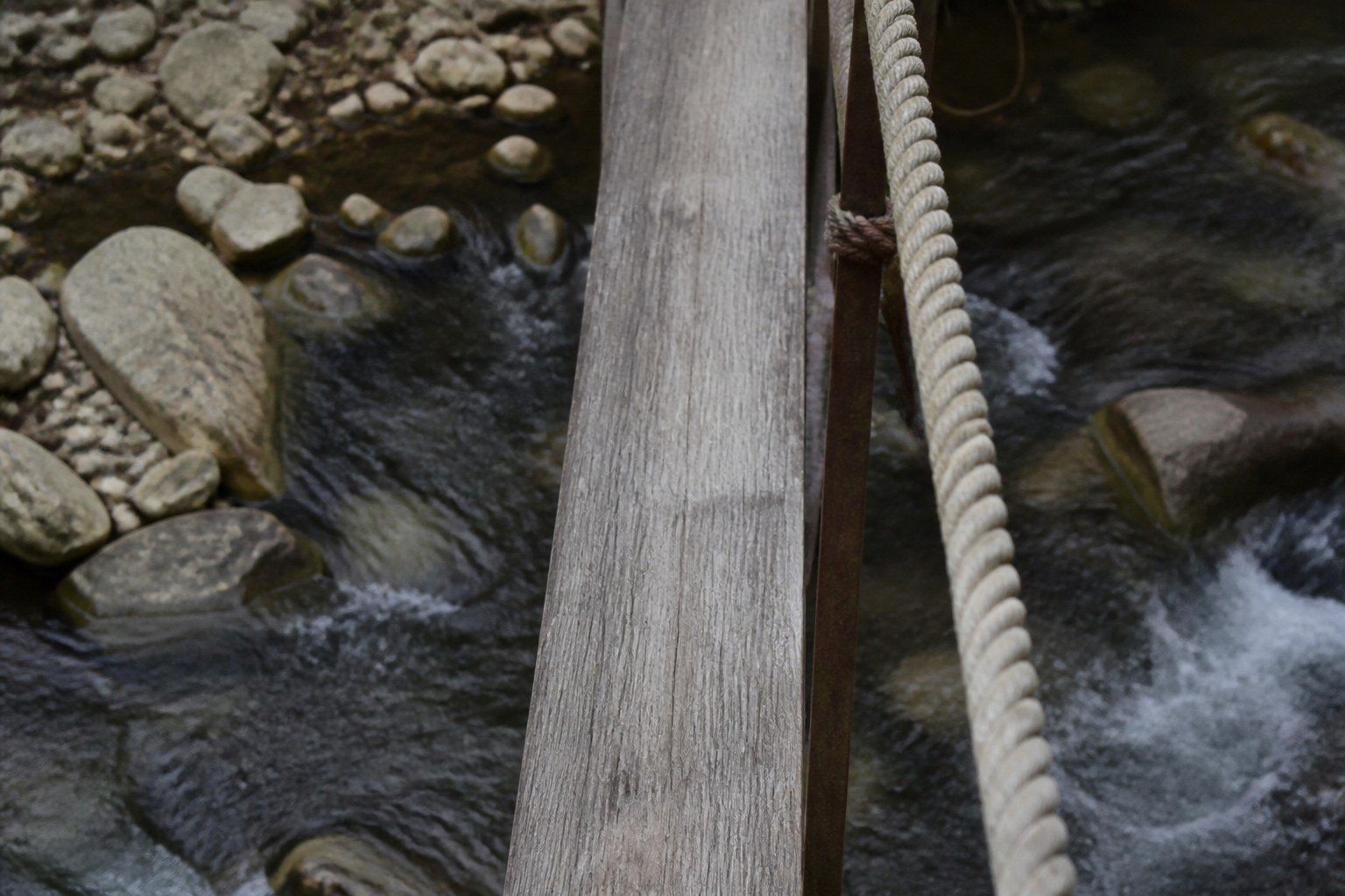 A slat of a rope bridge over a stream with a fixed handrail beside it