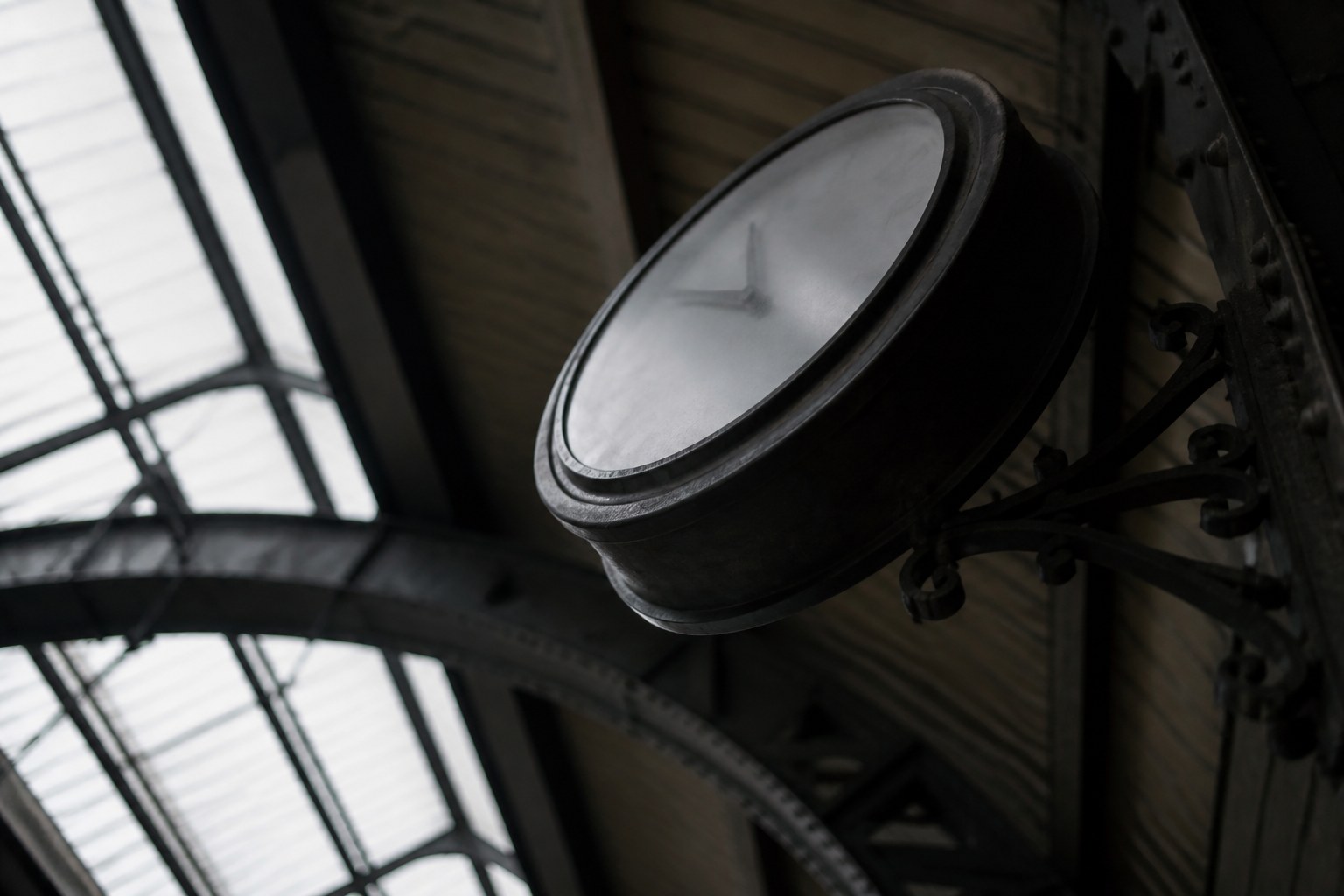A station platform clock seen from below at a steep angle so its face is foreshortened, iron canopy above
