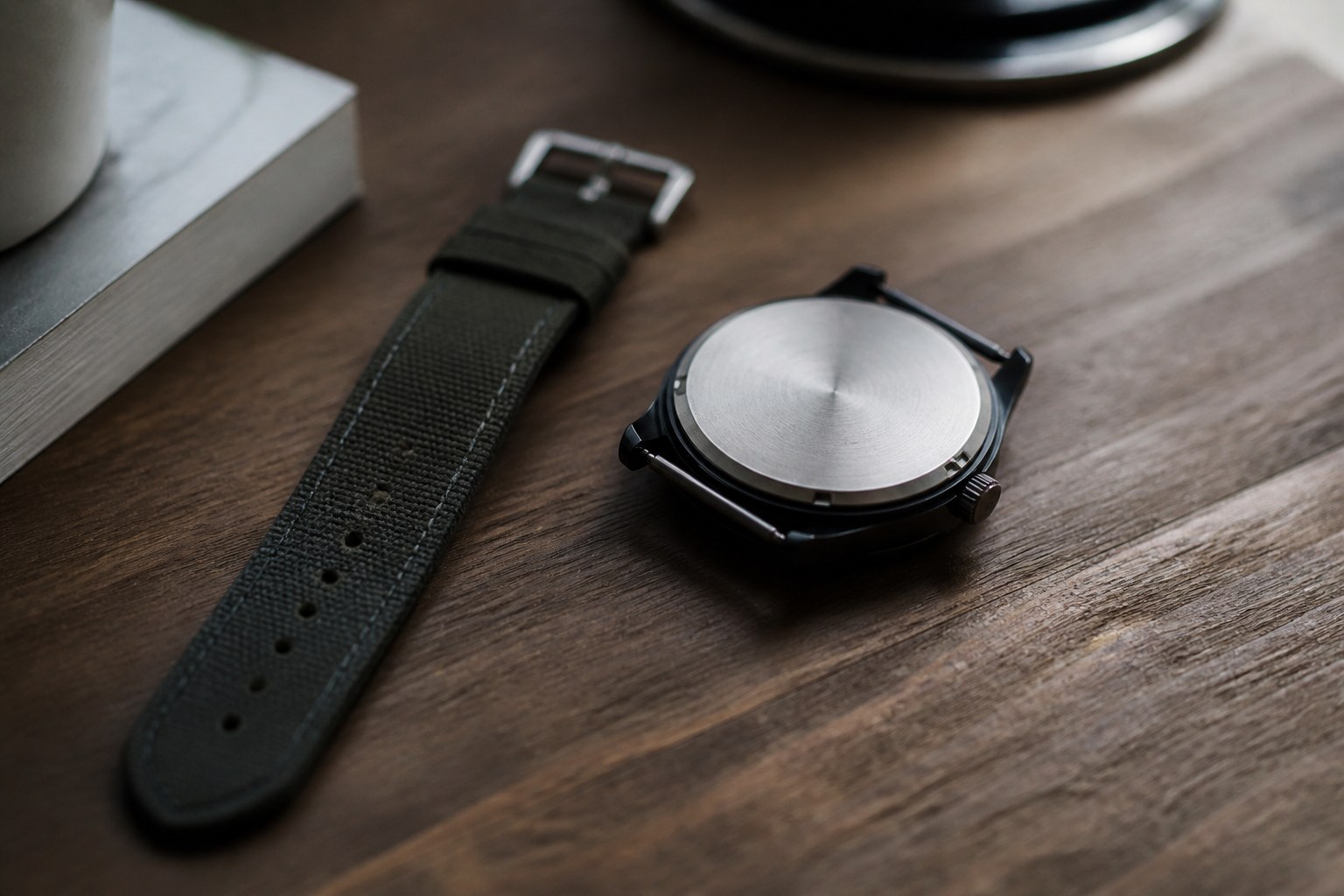 A wristwatch lying face down on a wooden desk beside its unbuckled strap