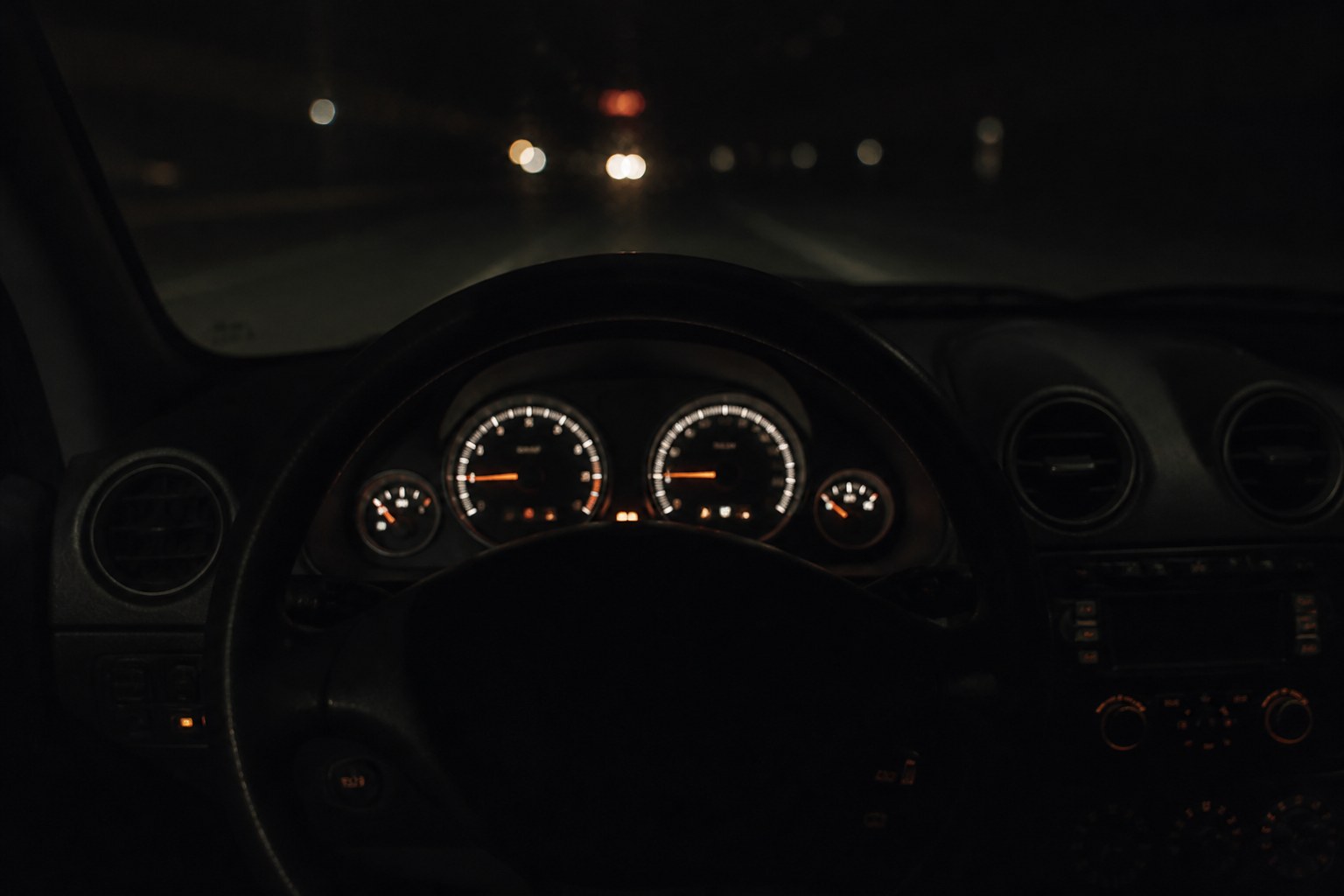A car dashboard at night with instrument light spilling across the wheel, the windscreen dark