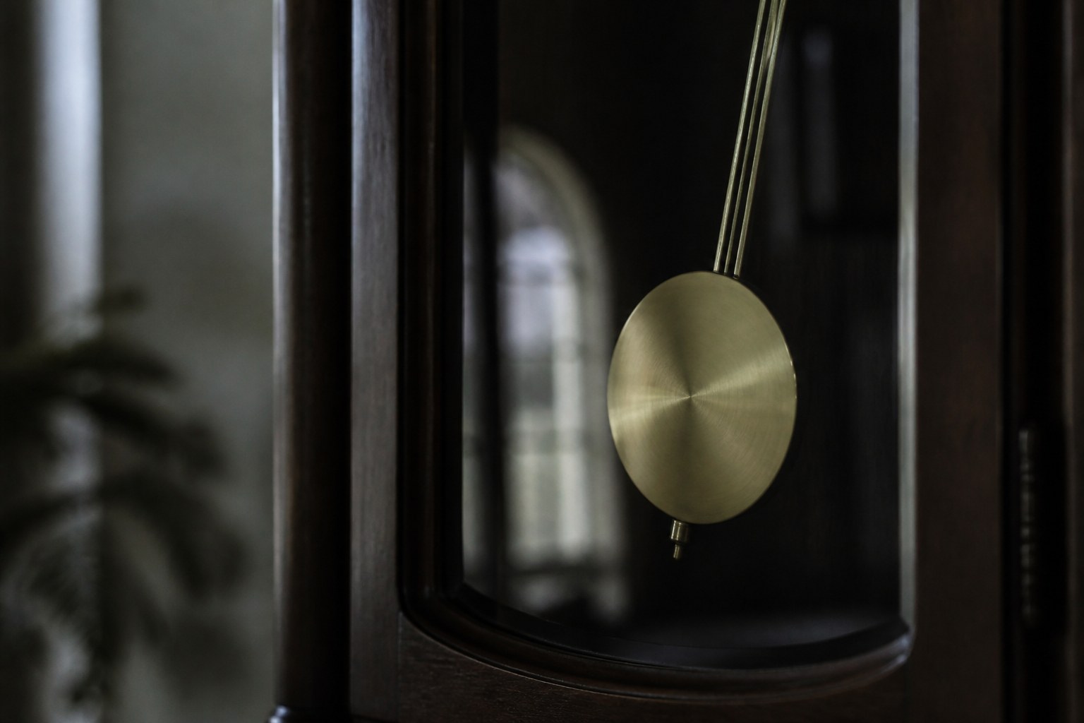 A grandfather clock pendulum caught mid swing behind its glass door, the case dark