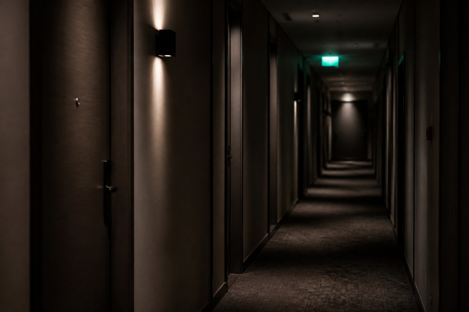 A hotel corridor at night with a lit exit sign and a row of closed doors