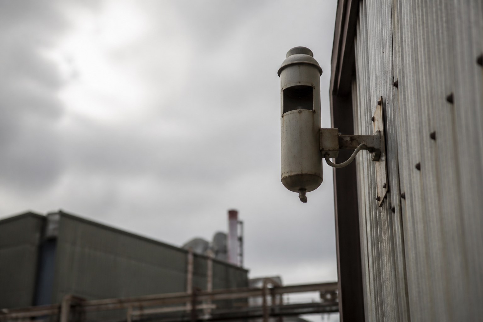 A shift whistle mounted high on a factory wall against an overcast sky