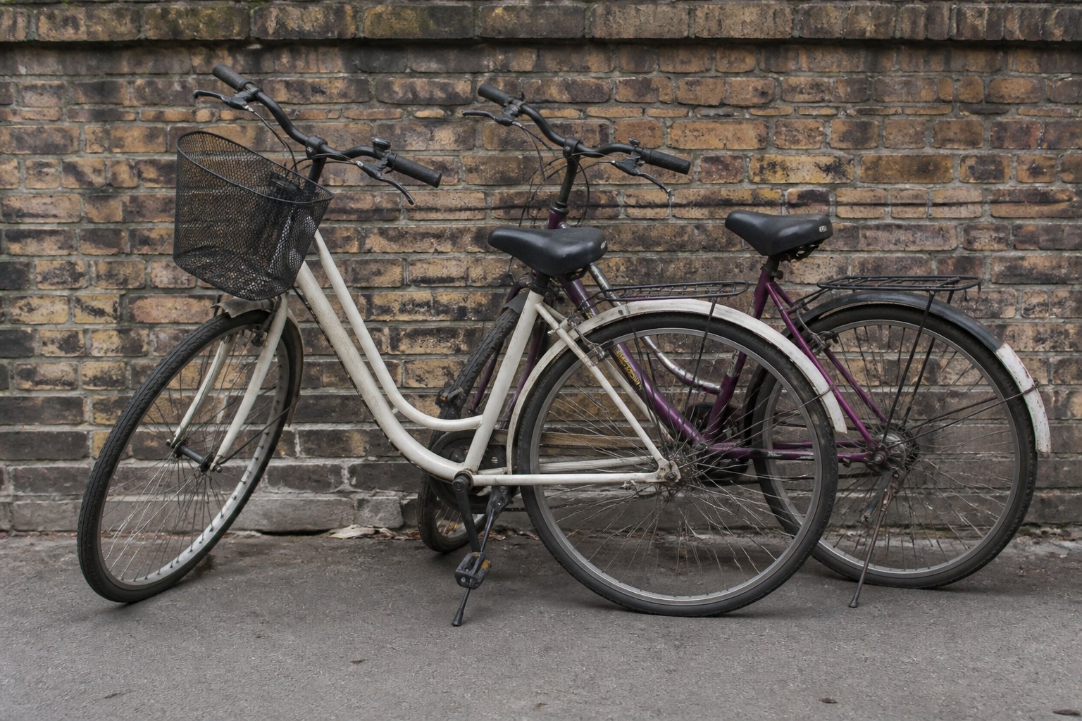 Two bicycles of different colours leaning together outside a brick wall