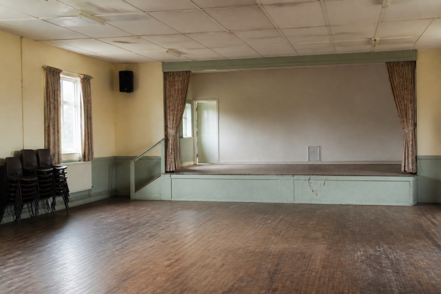 An empty village hall with a small stage at one end and stacked chairs