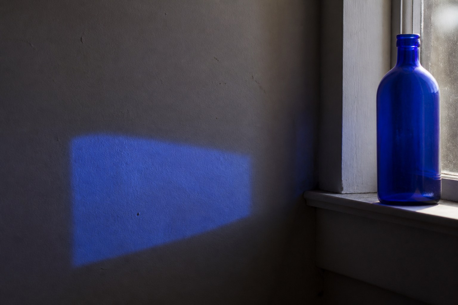 A rectangle of blue light cast on a grey interior wall, with a cobalt glass bottle standing on the windowsill beside it