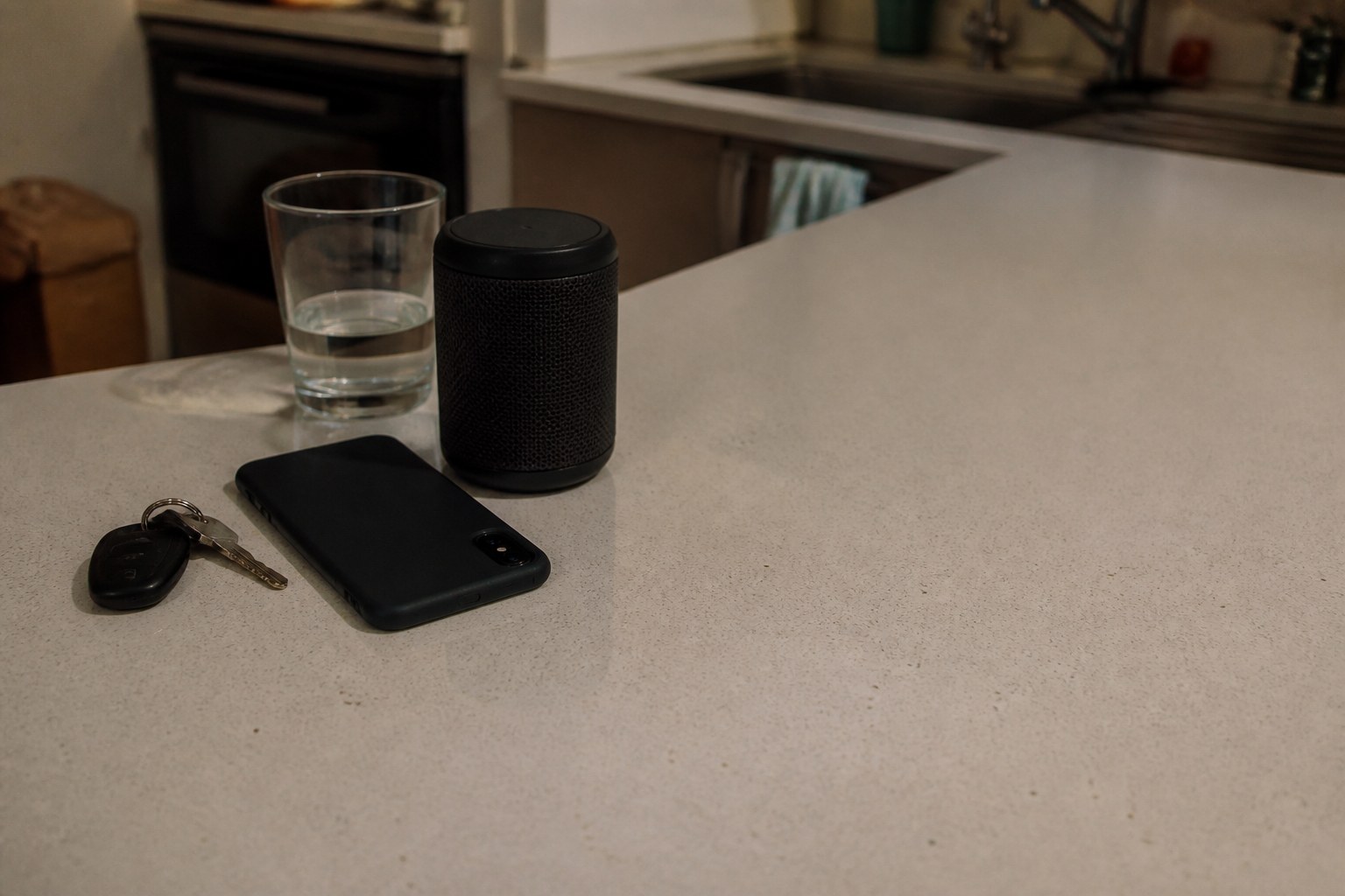 A kitchen counter at dusk with a glass of water, a small speaker, a phone lying face down, and a set of car keys