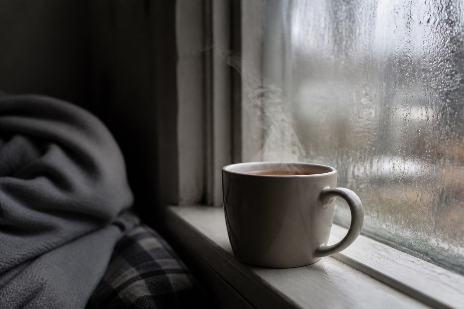 A steaming mug on a wooden windowsill beside a cold window pane running with condensation, a folded blanket on the chair behind