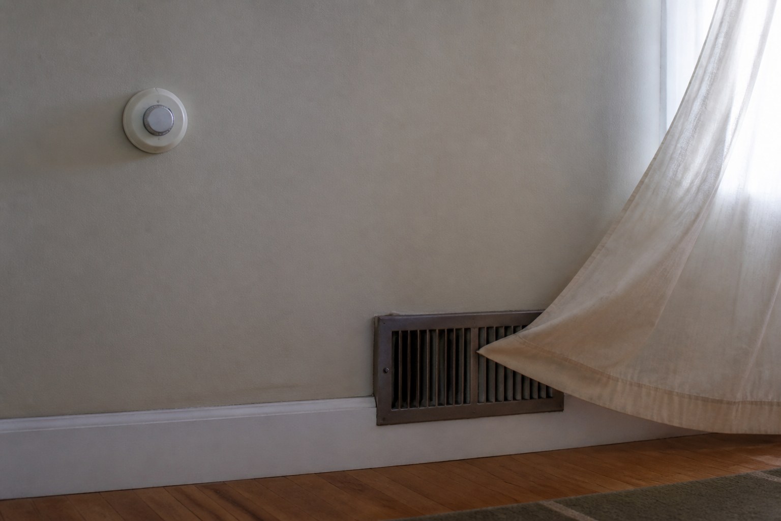 An interior wall with a round thermostat, a floor vent at the baseboard, and a sheer curtain lifting above it