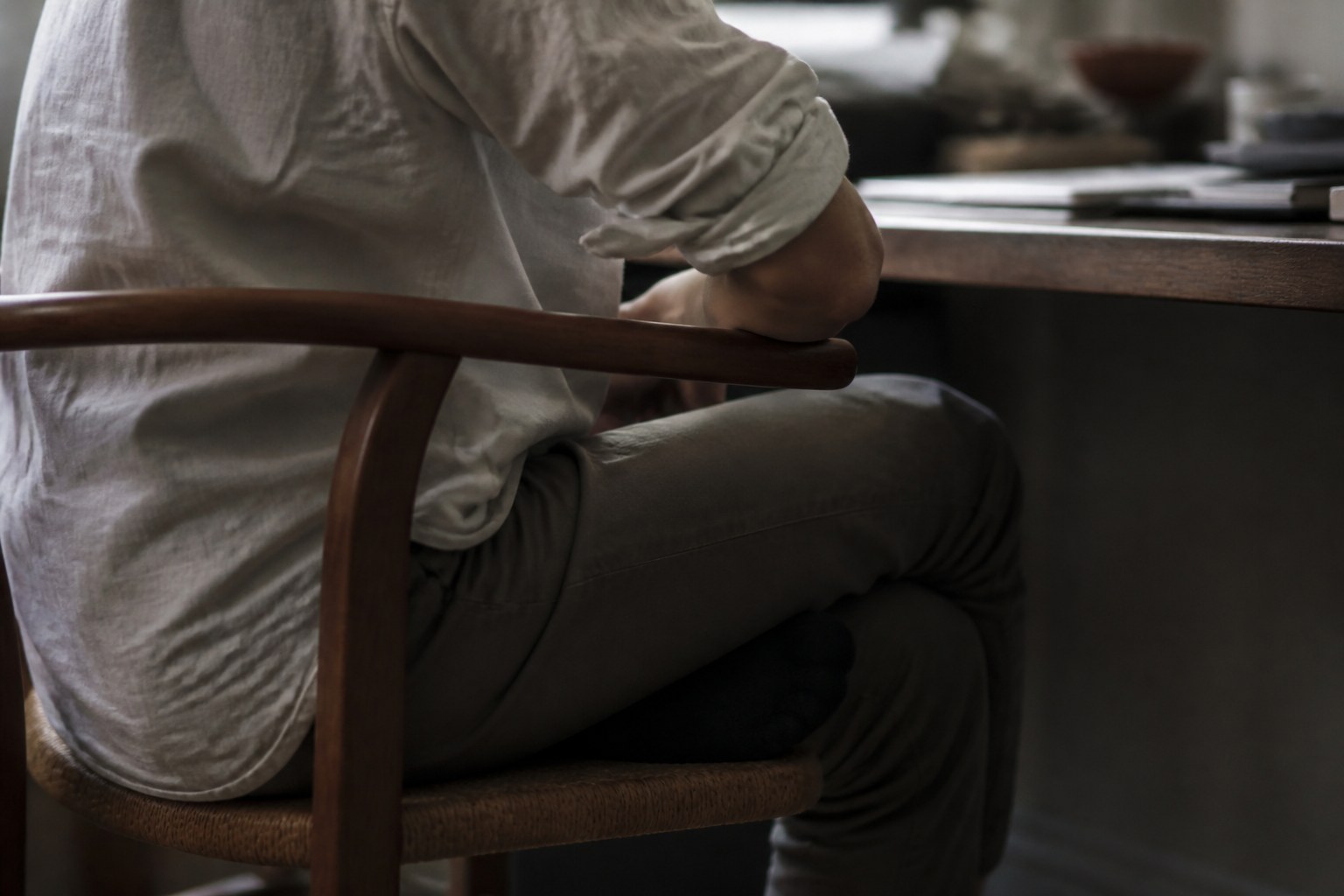 A person seated sideways at a desk with one leg crossed under the chair and a forearm pressed against the wooden chair arm, no face visible