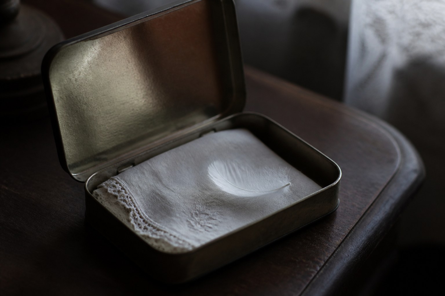 An open metal keepsake tin on a dark table holding a folded lace edged handkerchief and one small white feather