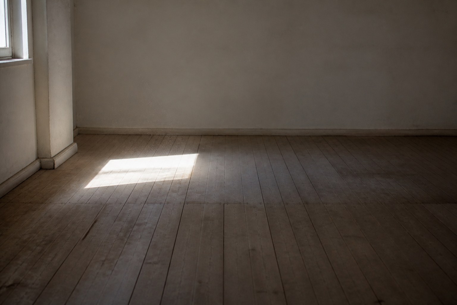 An empty room with bare plaster walls and wide floorboards, lit by a bright rectangle of sunlight from the window