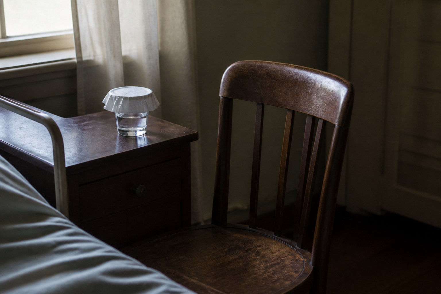A single wooden chair drawn close beside a bedside table holding a covered glass of water, the room otherwise empty