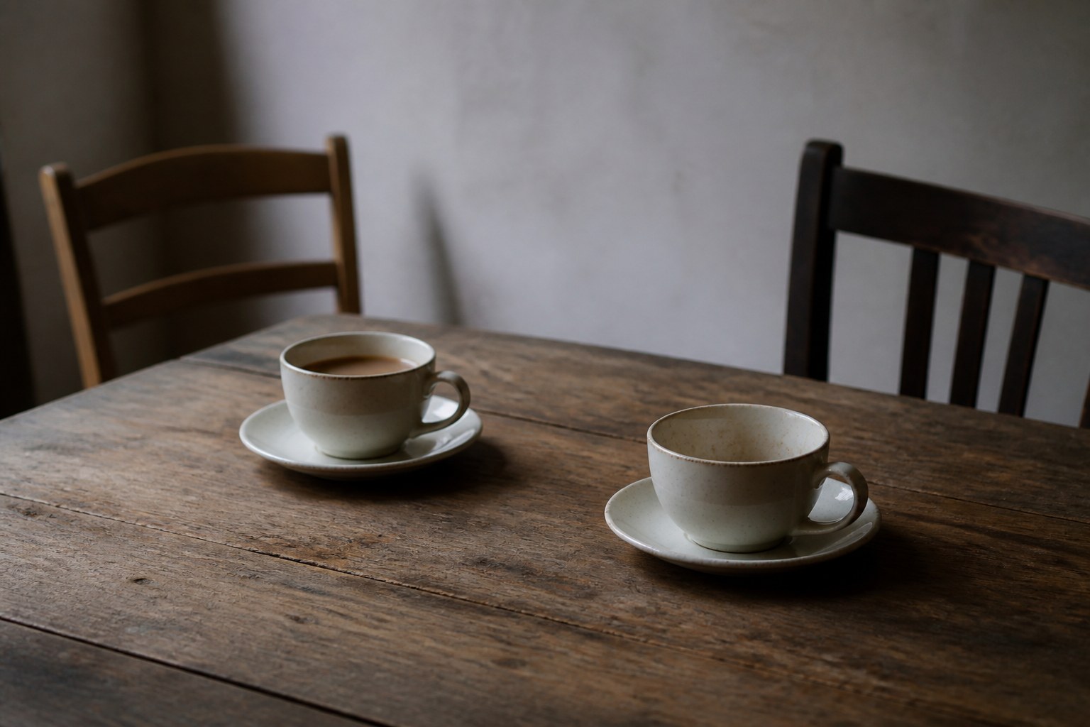 Two teacups on a bare wooden table with the chairs pushed back at different angles, one cup left untouched