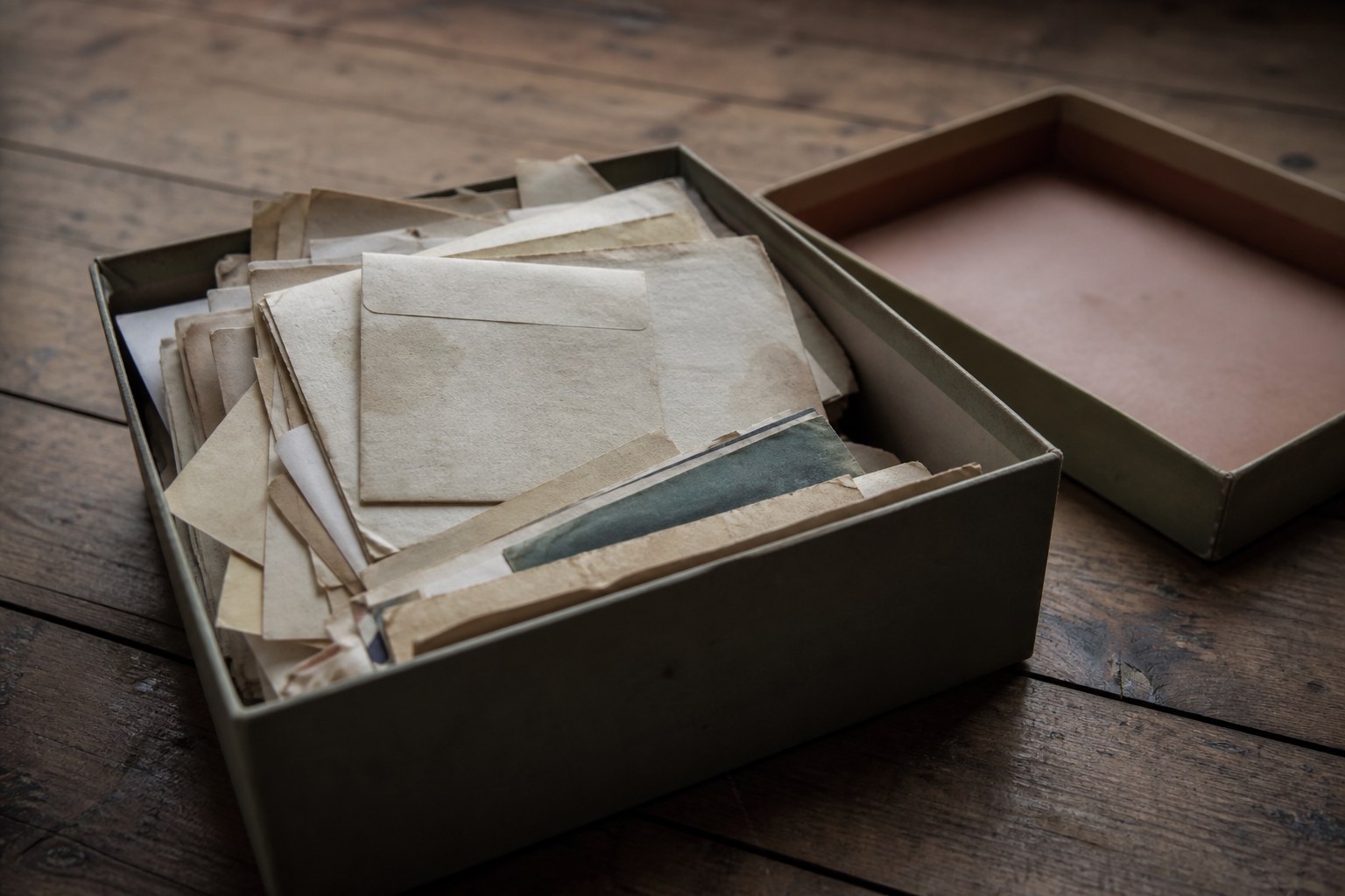 A shoebox with its lid set aside on bare floorboards, filled with loose old letters and papers