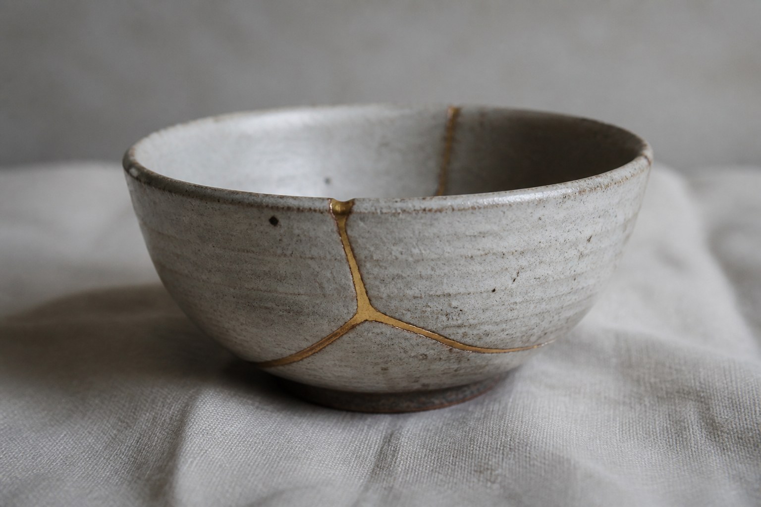 A ceramic bowl broken and repaired, the mended joins running visibly across it, resting on plain cloth