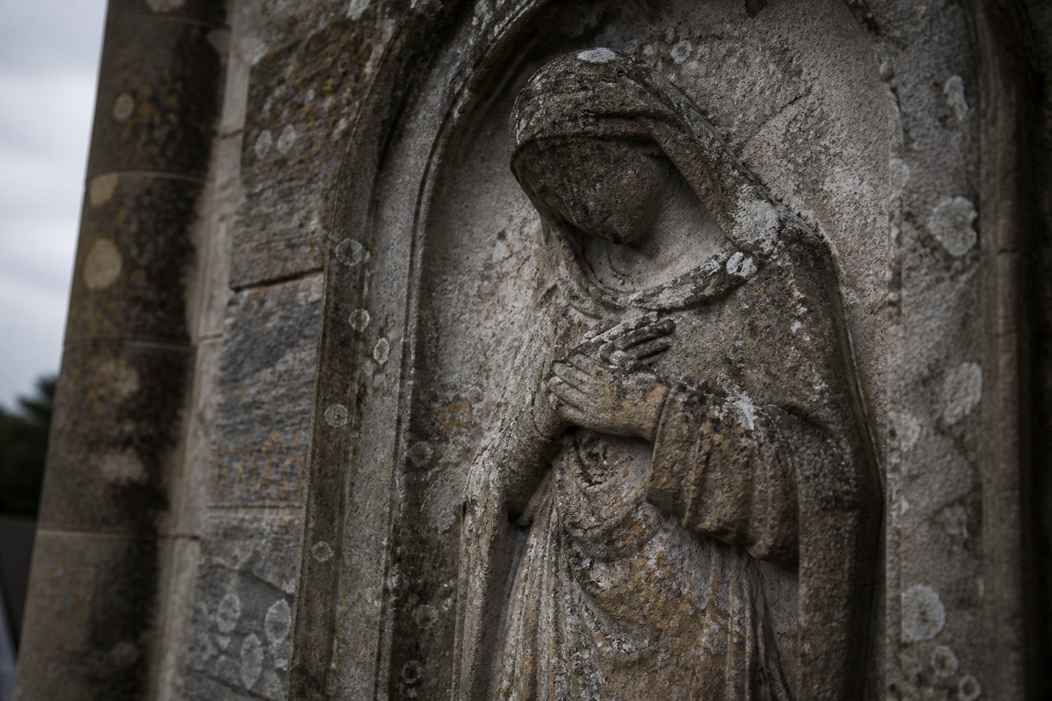 A weathered stone carving on a church wall showing a robed figure with a bowed head and hands drawn in at the chest, the surface worn and spotted with lichen