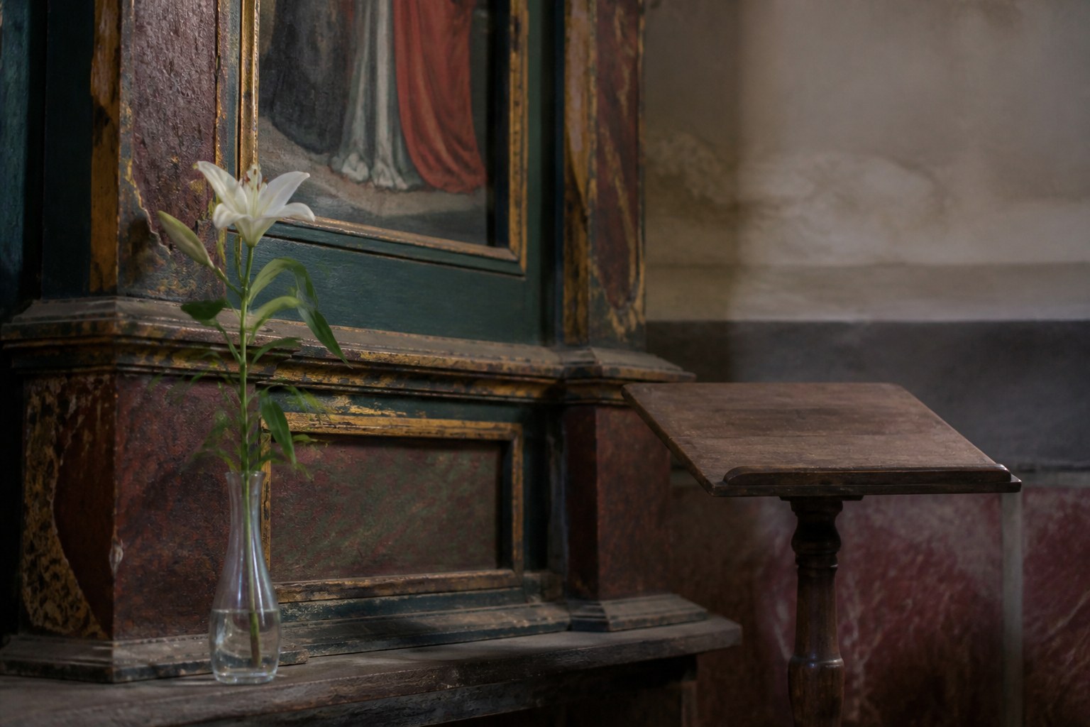 The corner of an old painted altar panel with a single white lily standing in a clear glass vessel and a small wooden reading desk beside it