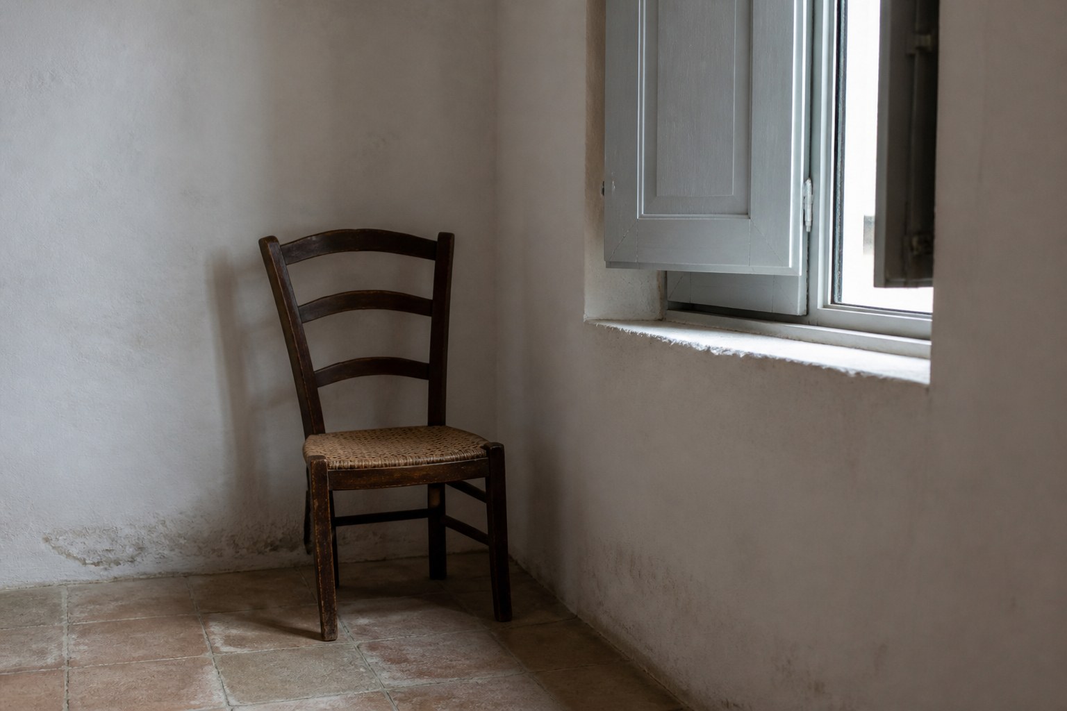 A plain wooden chair beside a shuttered window in a bare whitewashed room