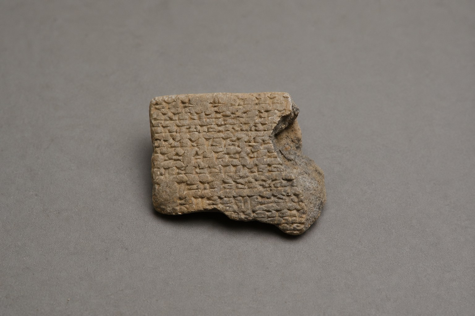 A small broken fragment of a clay tablet impressed with rows of wedge-shaped cuneiform marks, photographed on a plain grey surface